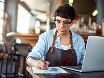 Female small business owner reviewing documents and taking notes with laptop nearby