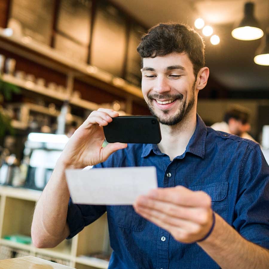 A smiling man takes a picture with his smart phone of a check or paycheck for digital electronic depositing, also known as "Remote Deposit Capture"