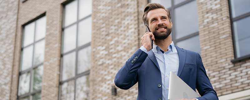 Businessman wearing stylish suit talking on mobile phone, holding laptop standing on the street. Successful business