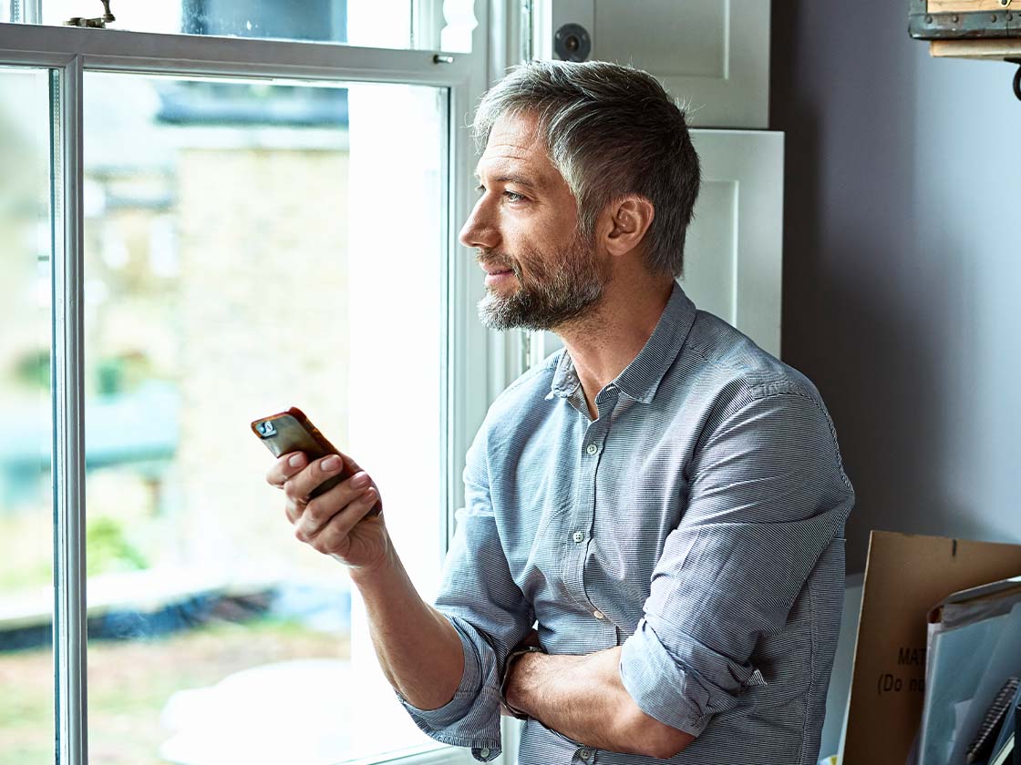 Mature businessman using phone in home office looking through window